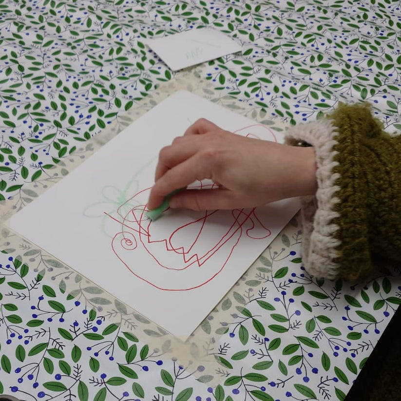A person's hand drawing on a piece of paper with green chalk, placed on a white table covered in a green and purple leaf patterned tablecloth.