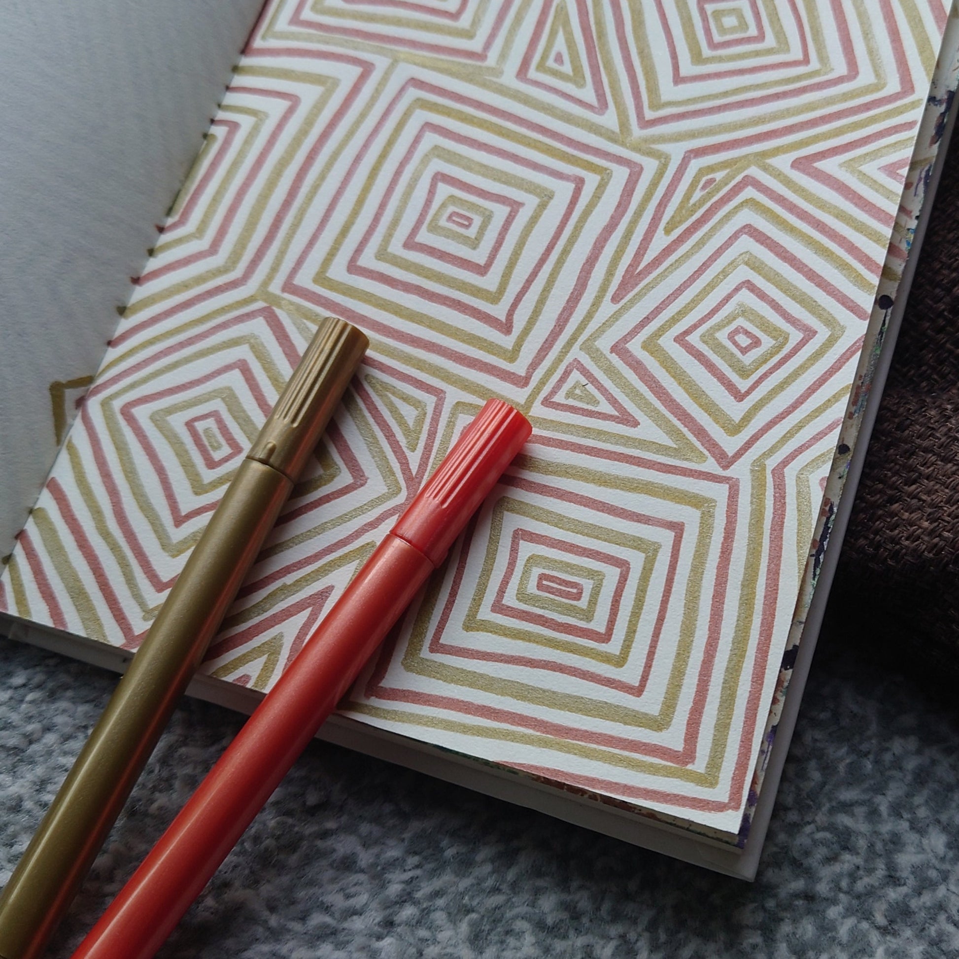 A notebook with a pink, white, and green geometric diamond pattern, accompanied by two pens (one red, one gold) resting on top, set against a gray textured fabric background.