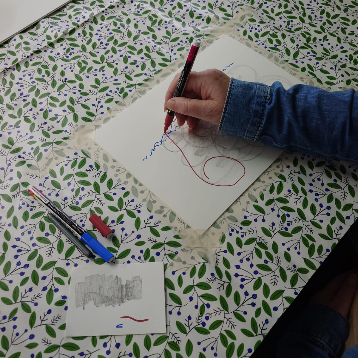 A person's hand drawing on a white sheet of paper with a red pen, placed on a green and purple leaf-patterned tablecloth alongside other colored pencils and a small sketch card.