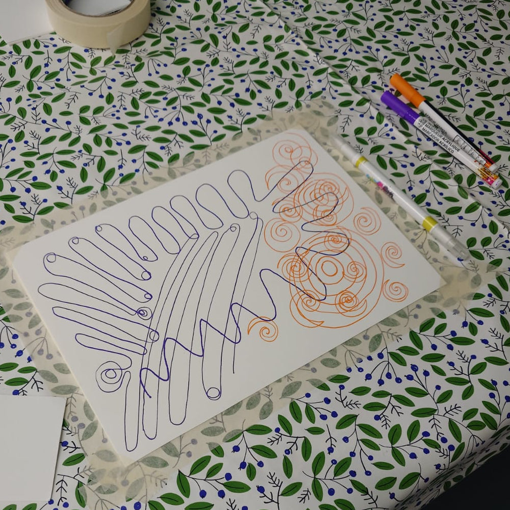 A white sheet of paper with abstract hand-drawn line art in blue and orange, placed on a patterned green and purple tablecloth with markers and tape nearby.