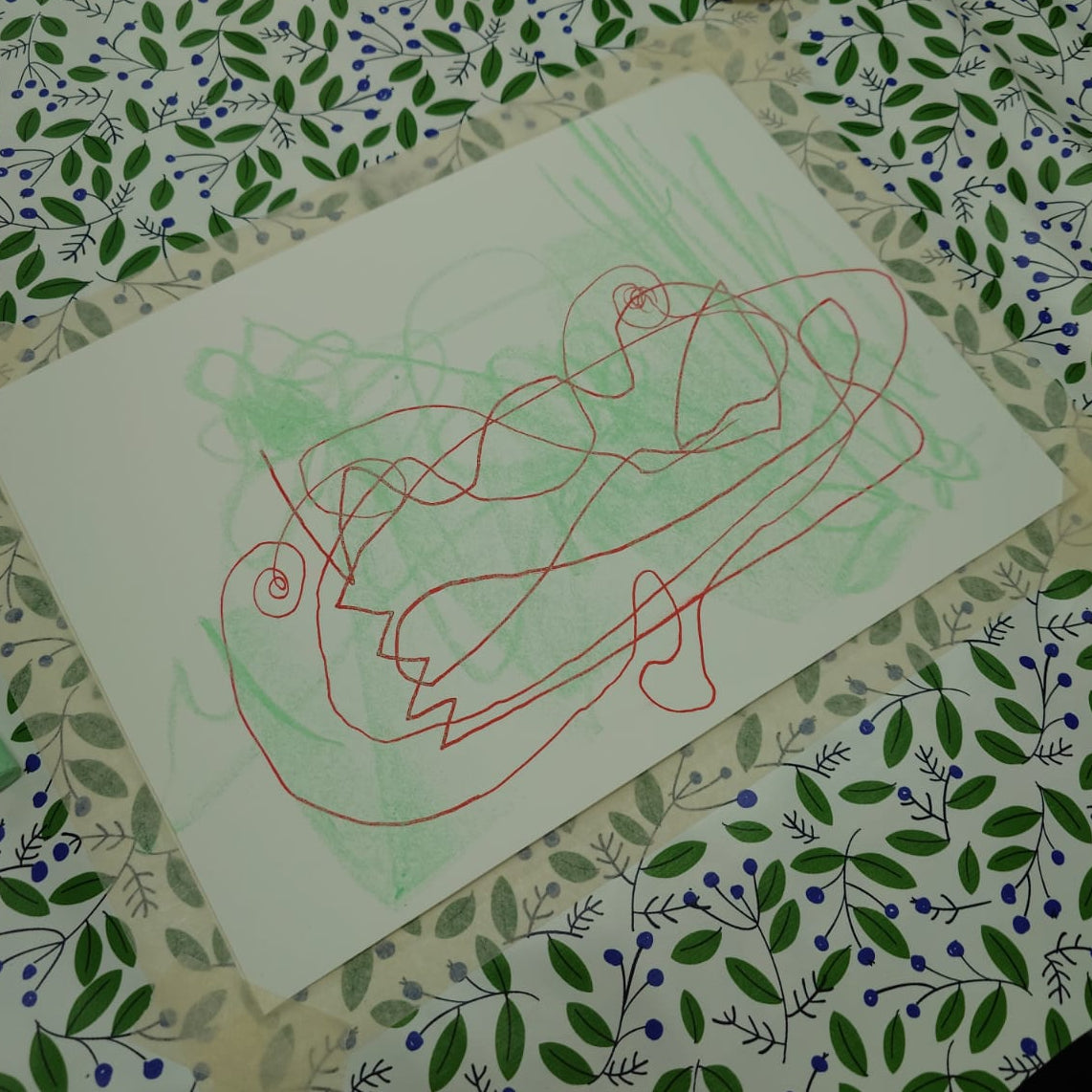 A white rectangular sheet of paper with abstract scribbled lines in red and light green, placed on a patterned surface with a small matching card in the upper right.