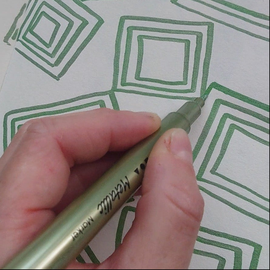 A hand holding a metallic green pen, drawing green geometric square patterns on a white sketchbook page.